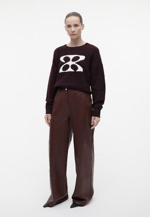 Bordeaux gekleurde gebreide trui met een wit geometrisch ontwerp, gecombineerd met hoog-waisted brede leren broek in bruin en puntige zwarte schoenen.