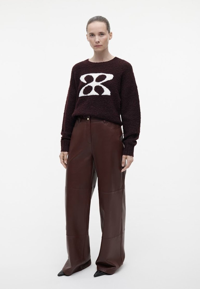Bordeaux gekleurde gebreide trui met een wit geometrisch ontwerp, gecombineerd met hoog-waisted brede leren broek in bruin en puntige zwarte schoenen.
