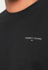 Μαύρη βαμβακερή μπλούζα με στρογγυλή λαιμόκοψη, με το λευκό κείμενο "TOMMY JEANS" και ένα μικρό πολύχρωμο λογότυπο από κάτω. Απλός, καθημερινός σχεδιασμός.