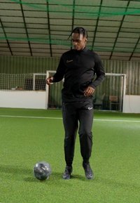Nike zwarte sportoutfit met lange mouwen en een lange broek, presterend op kunstgras, nabij een zwart en zilver voetbal.