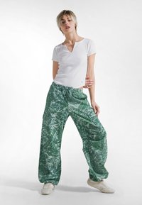 Bílá košile s krátkým rukávem spojená s volnými zelenými kalhotami s listovým vzorem. Sestavu doplňují světle zbarvené boty.