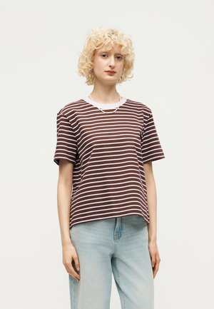 Persoană tânără cu păr blond creț, purtând un tricou maro și alb cu dungi și blugi albastru deschis pe un fundal simplu.