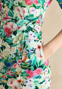 Blumenkleid mit einem hellgrünen Grundton, verziert mit einer Vielzahl bunter Blumen und Blätter. Knoten-Detail in der Taille und seitlicher Tasche.