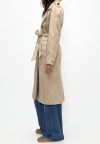 Kobieta ubrana w beżowy trencz z paskiem noszący szerokie dżinsy i zielone sneakersy, stojąca bokiem na tle białym.