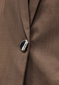 Καφέ ύφασμα με υφή υφαντού, που διαθέτει ασημί κουμπί με μαύρη λεπτομέρεια. Το ύφασμα είναι λείο και έχει ελαφριά γυαλάδα.