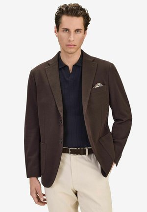 Man som bär en mörkbrun kavaj, marinblå pikétröja, beige byxor och en mönstrad näsduk i kavajfickan.