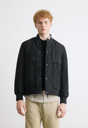 Joven con cabello rizado que lleva una chaqueta negra abotonada sobre una camisa a rayas y pantalones beige, de pie frente a un fondo blanco liso.