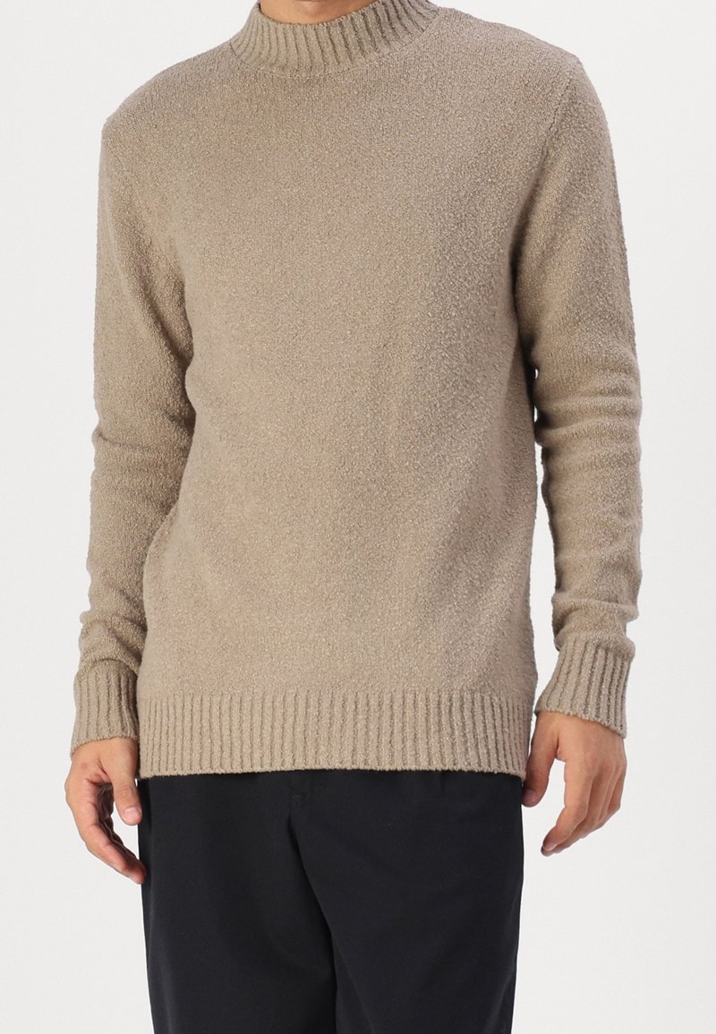 Mann, der einen beigen strukturierten Strickpullover mit gerippten Bündchen und Saum trägt, kombiniert mit lockeren schwarzen Hosen.