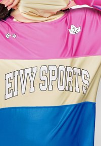 Haut de sport coloré avec une section supérieure rose, une section centrale crème portant "EIVY SPORTS" en lettres gras, et une section inférieure bleue.