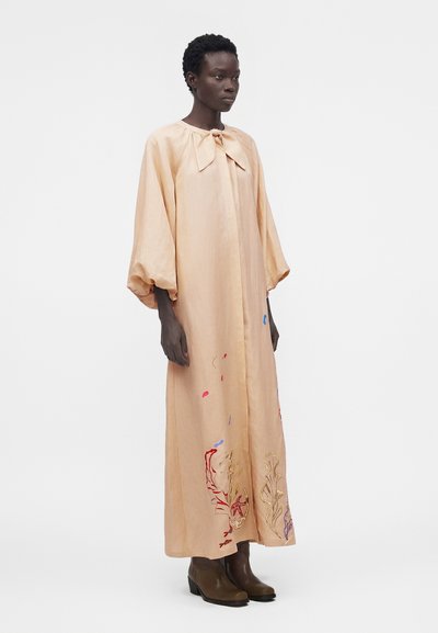 Γυναίκα που φοράει ένα άνετο, ανοιχτό ροδακινί μακρύ φόρεμα με φουσκωτά μανίκια και πολύχρωμη κεντητή floral διακόσμηση κοντά στο κάτω μέρος, συνδυασμένο με καφέ μποτάκια μέχρι τον αστράγαλο.