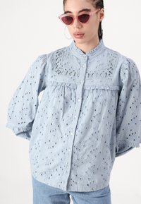 Light blue cotton blouse with puffed sleeves, embroidered eyelet design, ruffled neckline, and front button closure.