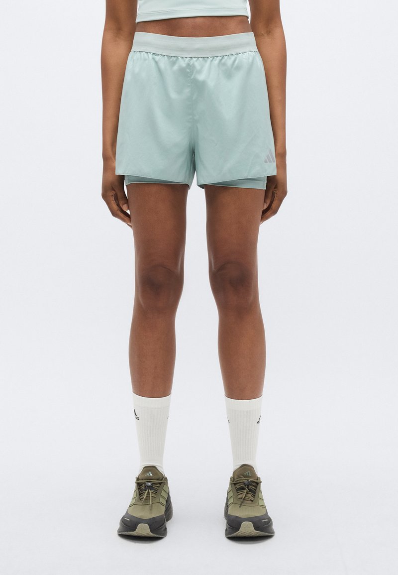 Shorts de sport vert clair avec une taille élastique, des fentes latérales et un logo discret. Associés à des chaussettes blanches montantes et des baskets olive.