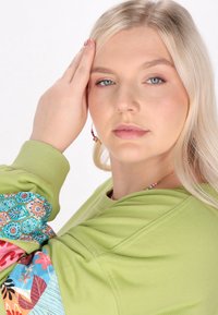 Felpa verde con maniche a fantasia multicolore; modella con capelli biondi di lunghezza media, che indossa orecchini a cerchio e una collana.