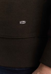 Brown ribbed sweater fabric with a round silver logo labeled "Petrol." Features a textured finish and contrasting stitching at the hem.