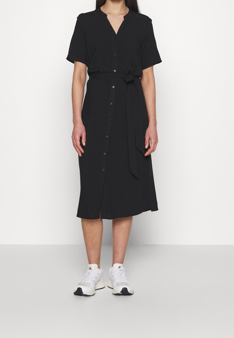 Robe noire à manches courtes avec un décolleté en V, des boutons à l'avant et une ceinture à la taille. Fabriquée dans un tissu doux avec une coupe décontractée et une longueur au genou. Portée avec des baskets blanches.