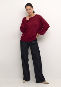 Burgundowa swetrowa dzianina o szerokich rękawach, zestawiona z ciemnoniebieskimi dżinsami i beżowymi butami na szpic. Prosty, casualowy i luźny fason.