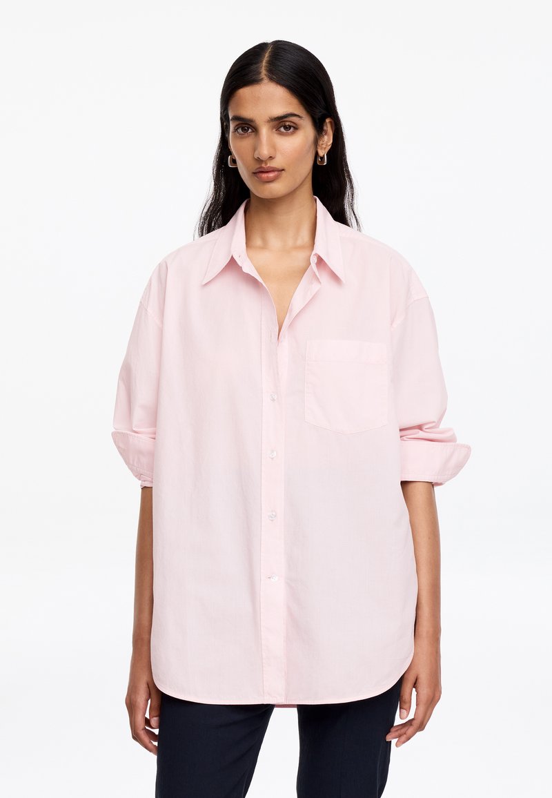 Femme aux longs cheveux foncés portant une chemise rose pâle surdimensionnée à boutons avec les manches retroussées et un pantalon noir, debout sur fond blanc.
