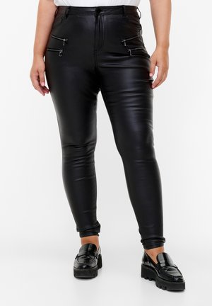 Schwarze Kunstlederhosen mit hoher Taille, schmalem Schnitt, Reißverschluss-Vordertaschen und glatter Textur. Kombiniert mit schwarzen Mokassins.