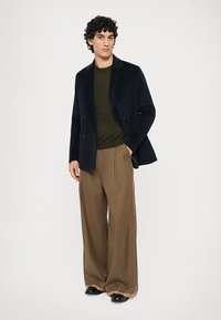 Man with curly hair wearing a black blazer, dark green sweater, wide brown pants, and black shoes, standing with one hand in pocket.