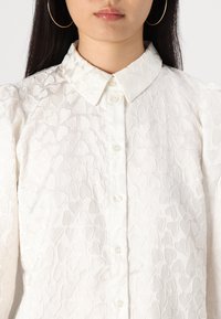 White blouse featuring a textured pattern of small hearts, a button-up front, and a classic collar design. Soft fabric with a subtle sheen.