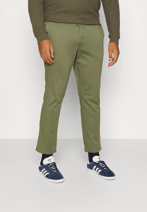 Mand iført olivengrønne bukser, mørke sokker og mørkeblå Adidas-sneakers, stående mod en ensfarvet hvid baggrund.