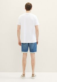 Camiseta blanca de mangas cortas, pantalones cortos de mezclilla doblados en el dobladillo, bolsillos traseros visibles, combinados con zapatillas de color claro, sobre un fondo neutro.