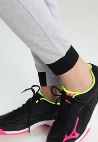 Pantalones de chándal grises con puños negros combinados con zapatillas deportivas negras que tienen acentos en verde neón y suelas rosas. Enfocados en la funcionalidad y la comodidad.