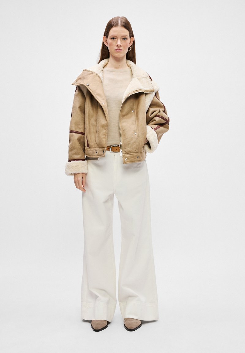 Braune Wildlederjacke mit einem cremefarbenen Lammfellkragen, weite cremefarbene Hose und beigem Schuhwerk. Die Jacke hat einen Knopfverschluss und kontrastierende Akzente.