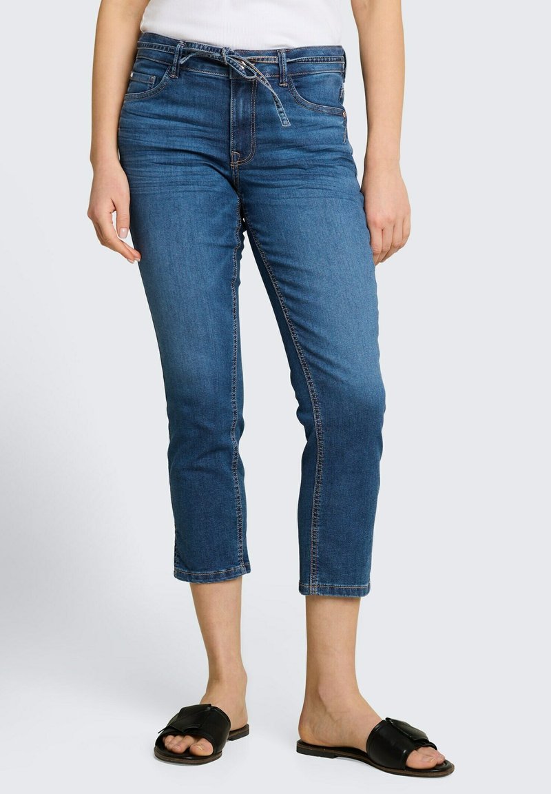 Femme portant un jean bleu mi-haut coupé avec une ceinture nouée en denim et des sandales noires à enfiler sur un fond clair.