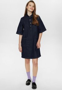 Donkerdenim button-up jurk met korte mouwen, twee voorzakken en een afgeronde zoom. Draag het met lavendelkleurige sokken en zwarte schoenen.