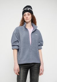 Γκρι φλις μπουφάν με ψηλό γιακά και τσέπες με φερμουάρ, συνδυασμένο με ροζ πουκάμισο από μέσα. Μαύρος σκούφος με λευκές λεπτομέρειες.