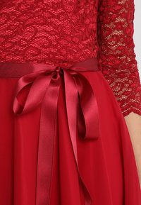 Gros plan d'une robe rouge avec un haut en dentelle, un ruban en satin noué en un nœud à la taille, et des manches en dentelle transparentes mi-longues.