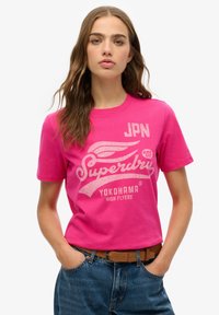 T-shirt en coton rose avec des graphismes imprimés blancs, incluant "Superdry", "Yokohama" et "JPN #01". Conception à manches courtes et col rond.