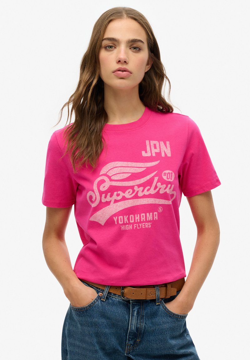 T-shirt en coton rose avec des graphismes imprimés blancs, incluant "Superdry", "Yokohama" et "JPN #01". Conception à manches courtes et col rond.