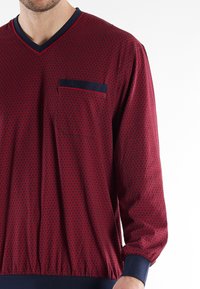 Hombre que lleva una camisa de manga larga de color rojo con un patrón, con ribetes azul marino en el cuello, el bolsillo del pecho, los puños y la cintura.