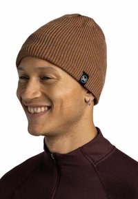 Brown ribbed knit beanie with a textured finish, featuring a small black logo label. Worn by a person in a dark red zip-up top.