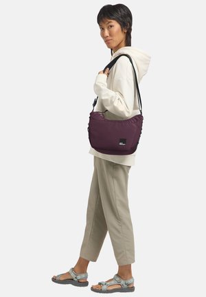 Sac en bandoulière violet rembourré avec une sangle noire et une marque. Porté avec un hoodie crème et un pantalon beige clair, se tenant contre un fond neutre.
