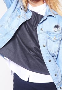 Giacca di jeans blu chiaro con dettagli strappati, indossata sopra una maglietta grigio ardesia e una camicia bianca con bottoni, con stratificazione visibile.