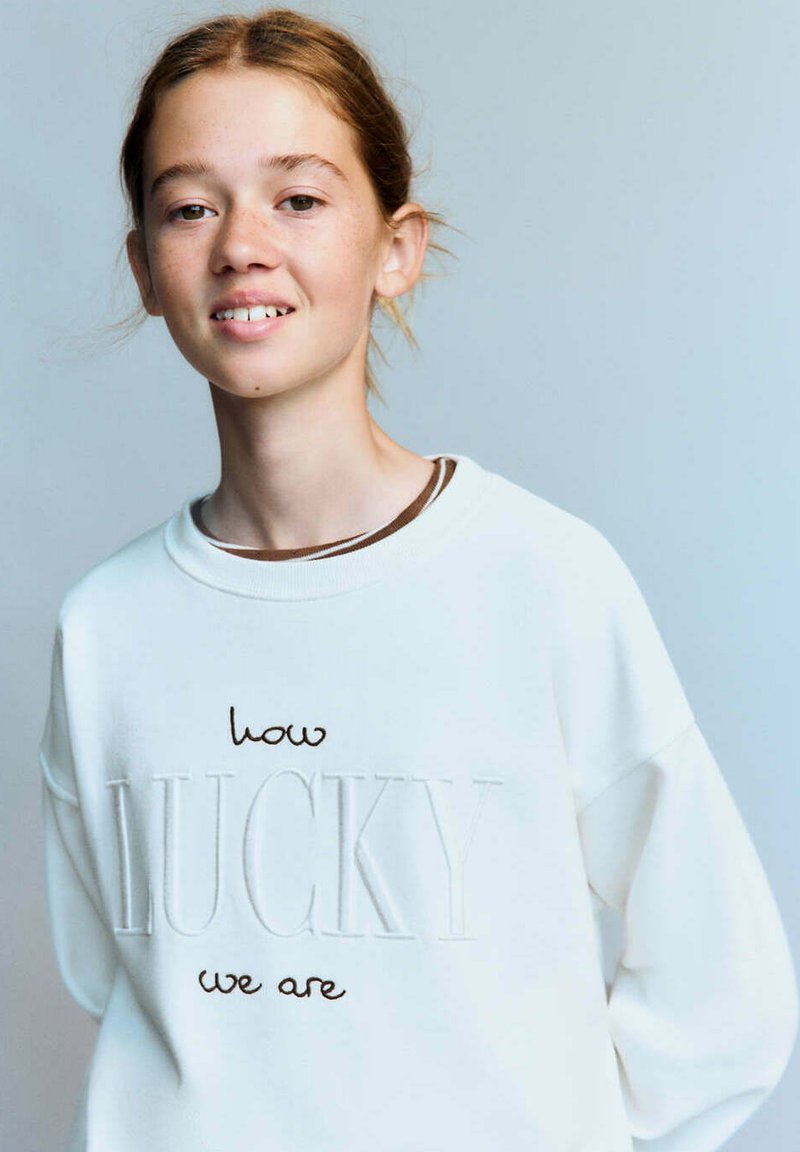 Jeune personne aux cheveux châtain clair attachés, portant un sweat-shirt blanc avec l’inscription "how LUCKY we are" sur un fond uni clair.