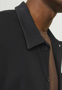 Black button-up shirt with a textured, ribbed design and a pointed collar, layered over a brown shirt. Features black buttons.