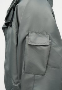 Γκρι αδιάβροχο μπουφάν με λεία υφή, φερμουάρ στη τσέπη του μανικιού και ρυθμιζόμενα κορδόνια στο κολάρο.