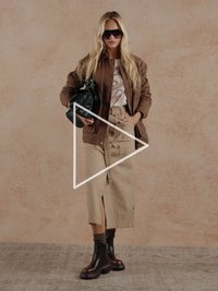 Bruine suède jas, grafisch T-shirt, beige midirok met een split, zwarte enkellaarsjes en oversized zonnebril. Houdt een zwarte handtas vast.