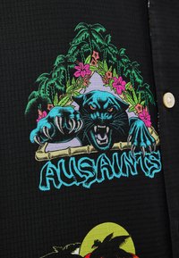 Camisa negra de manga corta con un gráfico colorido de un pantera, plantas tropicales y el texto "AUSSAINTS" en azul. Patrón de tela texturizada.