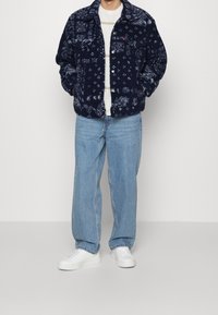 Giacca in pile blu navy con un motivo testurizzato, chiusura con bottoni e due tasche sul petto, abbinata a jeans azzurri e sneakers bianche.