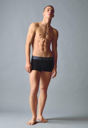 Ung mand står barfodet iført sorte, tætsiddende boxershorts og et sølvvedhæng, foran en ensfarvet grå baggrund.