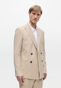Beige Doppelreiher-Anzug mit gespitztem Revers, strukturiertem Stoff, zwei Fronttaschen und braunen Knöpfen, kombiniert mit einem weißen Kragenhemd.