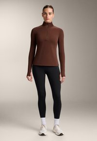 Top lung cu mânecă lungă de culoare maro, cu guler cu fermoar până la jumătate, asociat cu colanți negri. Proiectat cu mâneci ajustate și orificii pentru degete, realizat din material elastic.