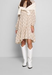 Beige plaid dress with long sleeves, ruffled hem, and a tied neckline. White knitted sweater worn over the shoulders. White cowboy boots.