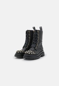 Botas de combate de cuero negro con tachuelas doradas en la puntera, cordones en la parte delantera con grandes ojales y una suela de goma gruesa y texturizada.