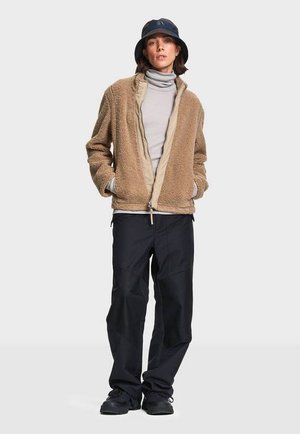 Frau trägt eine hellbraune Fleecejacke über einem grauen Rollkragenpullover, lockere schwarze Hose, schwarze Schuhe und einen dunklen Fischerhut, steht mit den Händen in den Taschen.